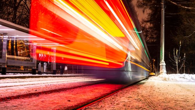 Public transportation in a city. Tram leaving the station, traffic lights, people, street, houses, overhead wires, rails. Movement and lights, long exposure.