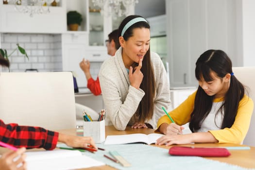 Asian mother helping daughter with school work, with brother in foreground and father in background. busy family working together at home in their kitchen.