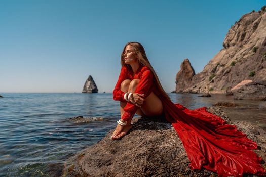 Beautiful sensual woman in a flying red dress and long hair, sitting on a rock above the beautiful sea in a large bay