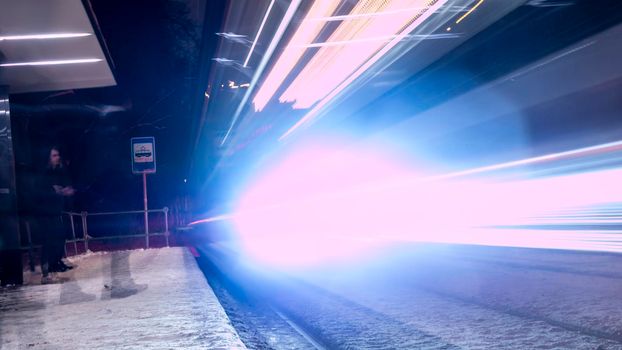 Public transportation in a city. Tram leaving the station, traffic lights, people, street, houses, overhead wires, rails. Movement and lights, long exposure.