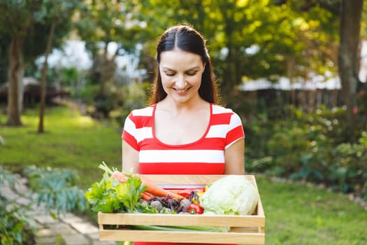 Smiling caucasian woman standing in garden holding box of fresh organic vegetables. gardening, self sufficiency and growing home produce.