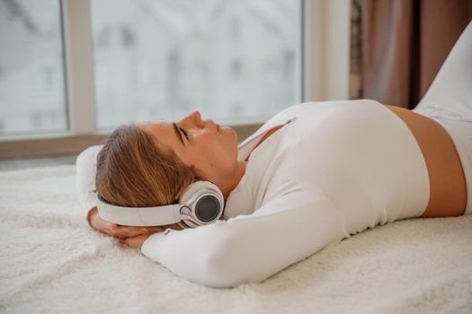 Side view portrait of relaxed woman listening to music with headphones lying on carpet at home. She is dressed in a white tracksuit