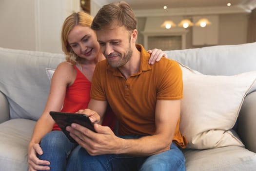 Caucasian couple sitting on couch smiling and using tablet at home. enjoying free time relaxing with technology at home.