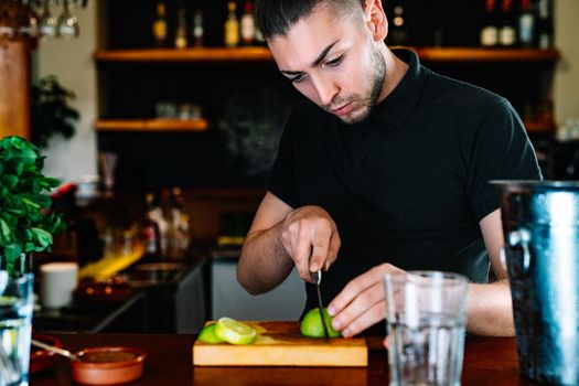 Young and modern waiter, with long dark hair, dressed in black polo shirt, cutting slices of citrus fruit on a wooden board, to prepare a cocktail. Waiter preparing a cocktail. Cocktail glass with ice cubes. Gin and tonic. Bar full of cocktail ingredients. Dark background and dramatic lighting.