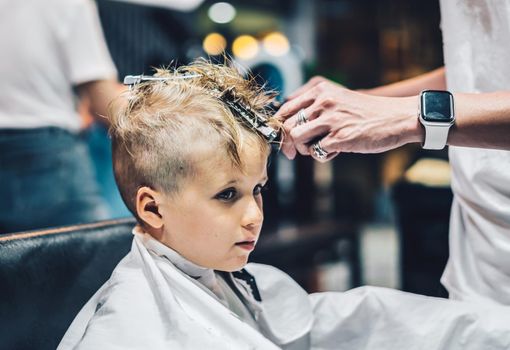 Lifestyle photo Barber shop blond boy customer. Hairdresser man work move hand comb tool scissors dryer clip white cape. Modern style kid short hair cut. Fashion business service for children.