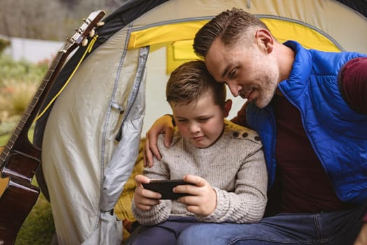 Caucasian father and son using smartphone while sitting in a tent in the garden. fatherhood and love concept