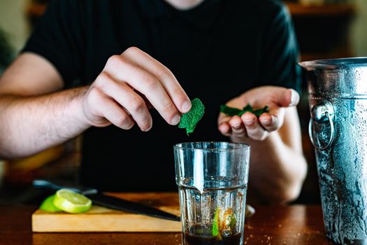 Young and modern waiter, adding mint in a large glass in the preparation of a mojito Waiter preparing a cocktail. Cocktail glass with ice cubes. Mojito. Bar full of cocktail ingredients. Dark background and dramatic lighting.