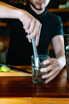 Young and modern waiter, with long dark hair, dressed in black polo shirt, crushing the ingredients of the mojito to release its juices. Waiter preparing a cocktail. Cocktail glass with ice cubes. Mojito. Bar full of cocktail ingredients. Dark background and dramatic lighting. Vertical