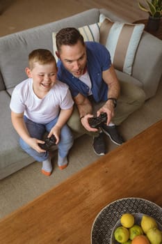 Overhead view of caucasian father and son playing video games sitting on the couch at home. gaming and entertainment concept