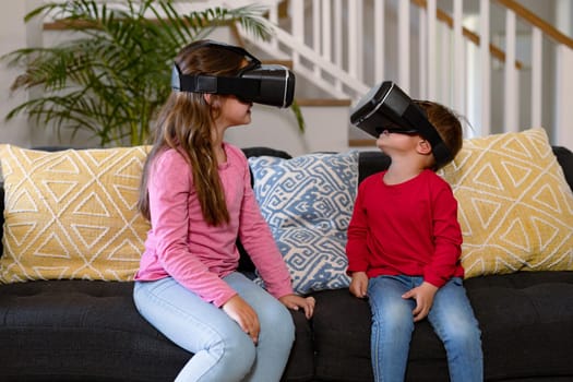 Happy caucasian siblings sitting on sofa, wearing vr headset and having fun. childhood, leisure and discovery using technology at home.