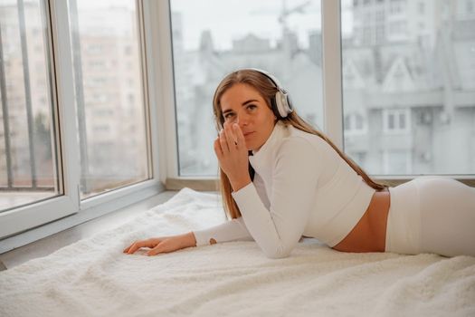 Side view portrait of relaxed woman listening to music with headphones lying on carpet at home. She is dressed in a white tracksuit