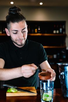 Young and modern waiter, with long dark hair, dressed in black polo shirt, adding brown sugar in a large crystal glass to prepare a cocktail. Waiter preparing a cocktail. Cocktail glass with ice cubes. Mojito. Bar full of cocktail ingredients. Dark background and dramatic lighting. Vertical