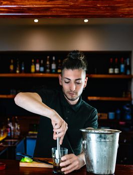 Young and modern waiter, with long dark hair, dressed in black polo shirt, crushing the ingredients of the mojito to release its juices. Waiter preparing a cocktail. Cocktail glass with ice cubes. Mojito. Bar full of cocktail ingredients. Dark background and dramatic lighting. Vertical