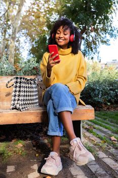 African American woman sit on park bench wearing headphones using mobile phone. Vertical image. Lifestyle concept.