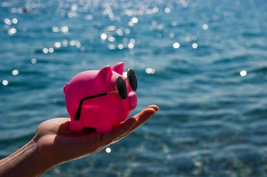 A woman holds a piggy bank in sunglasses on the background of the sea. Budget vacation