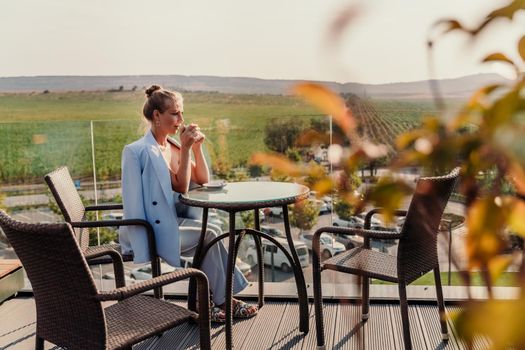 A middle-aged woman sits in a street cafe overlooking the mountains at sunset. She is dressed in a blue jacket and drinks coffee admiring the nature. Travel and vacation concept