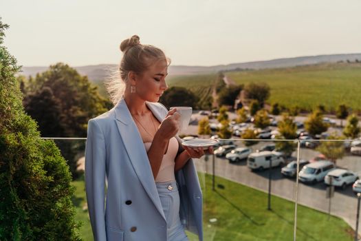 A middle-aged woman stands in a street cafe overlooking the mountains at sunset. She is wearing a blue jacket and drinking coffee while admiring nature. Travel and vacation concept