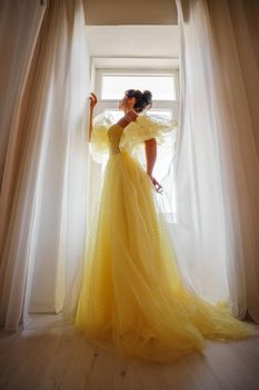 A woman's silhouette in a golden luxurious dress against the background of a window holds a curtain with her hands. Elegant lady in a yellow long silk dress with bare back, back view