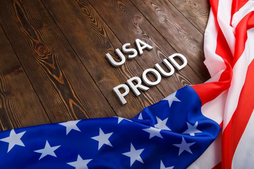 words USA proud laid with silver metal letters on brown wooden surface with flag of United States of America in diagonal view.