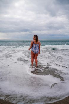 A plump woman in a bathing suit enters the water during the surf. Alone on the beach, Gray sky in the clouds, swimming in winter