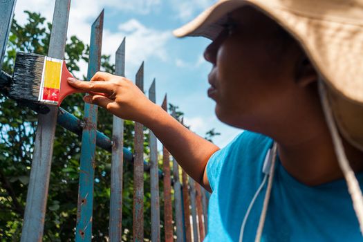 Latin young woman wearing a sun hat painting a fence with a brush in hands outdoors.