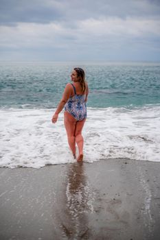 A plump woman in a bathing suit enters the water during the surf. Alone on the beach, Gray sky in the clouds, swimming in winter