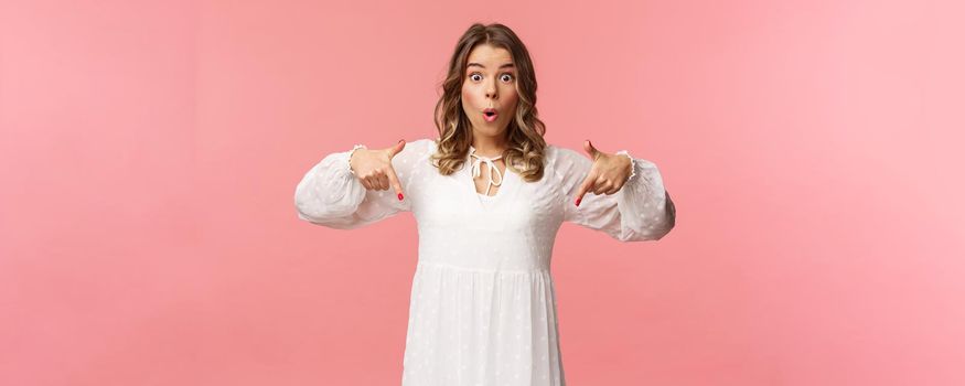 Waist-up portrait of interested, excited attractive blond 20s woman showing person something really cool, say wow pop eyes camera excited, pointing fingers down at great offer, pink background.