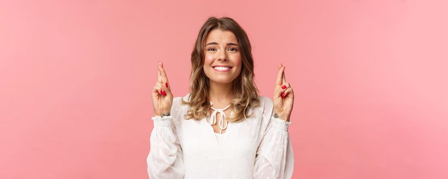 Close-up portrait of hopeful beautiful blond girl making wish, anticipating important results, cross fingers good luck and smiling as feeling slightly nervous, wear white dress, pink background.