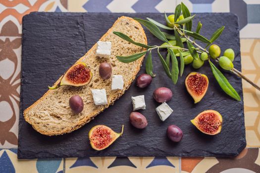 Green and black olives with loaf of fresh bread, feta cheese and young olives branch on olive wood chopping board over dark background.