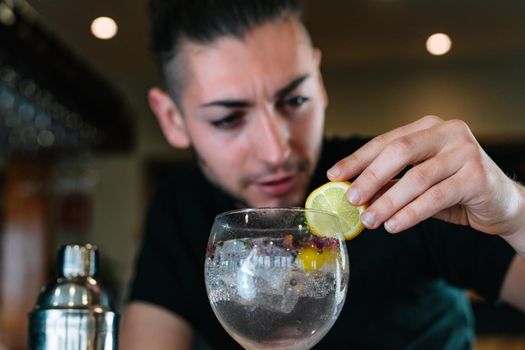 Young and modern waiter, with long dark hair, dressed in black polo shirt, decorating with a slice of citrus fruit a cocktail in a crystal glass with ice for a cocktail. Waiter preparing a cocktail. Cocktail glass with ice cubes. Gin and tonic. Bar full of cocktail ingredients. Dark background and dramatic lighting.
