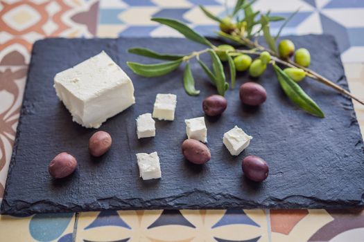 Green and black olives with loaf of fresh bread, feta cheese and young olives branch on olive wood chopping board over dark background.