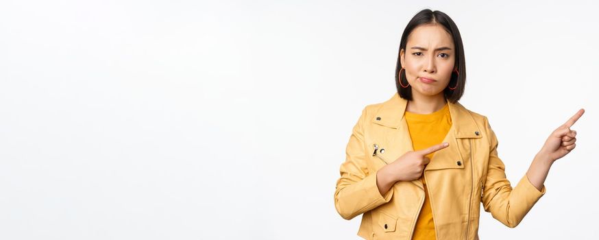 Disappointed asian girl frowning, sulking upset, pointing fingers right at advertisement, standing over white background. Copy space