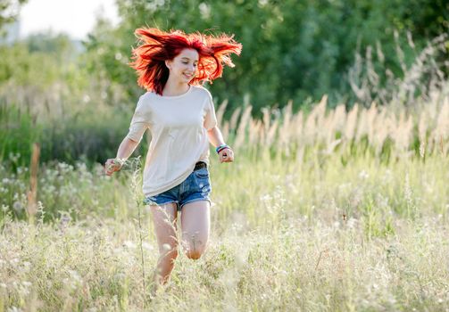 Teen girl with red hair running at the nature and smiling. Pretty young female in the sunny field