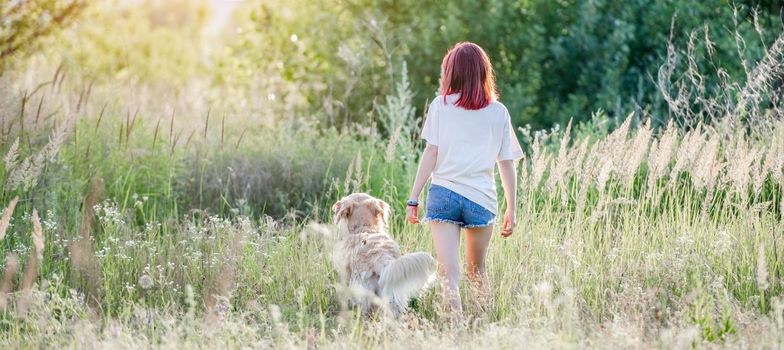 Girl with red hair walking with golden retriever dog at the nature. Back of pretty young female with doggy pet in the sunny field