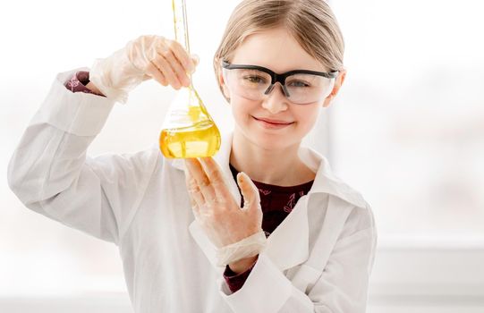 Smart girl during scientific chemistry experiment wearing protection glasses, holding bottle with orange liquid. Schoolgirl with chemical equipment on school lesson portrait