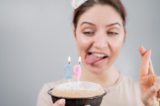 Happy caucasian woman sticking out her tongue and blowing out the candles on the cake with her fingers crossed. The girl celebrates her 29th birthday.