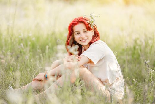 Girl with red hair sitting at the nature and smiling. Pretty young female in the sunny field with flowers