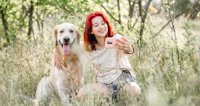 Teen girl with red hair with golden retriever dog making selfie on smartphone at the nature. Pretty young female with doggy pet and cell in the sunny field