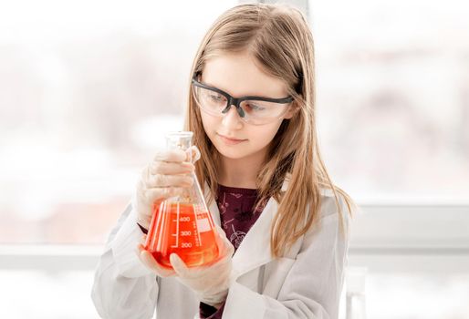 Smart girl during scientific chemistry experiment wearing protection glasses, holding bottle with orange liquid. Schoolgirl with chemical equipment on school lesson portrait