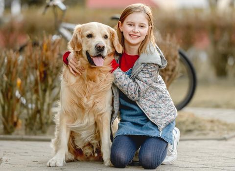 Preteen girl with golden retriever dog sitting in park in beautiful spring autumn day. Adorable female child kid hugging purebred doggy pet outdoors portrait