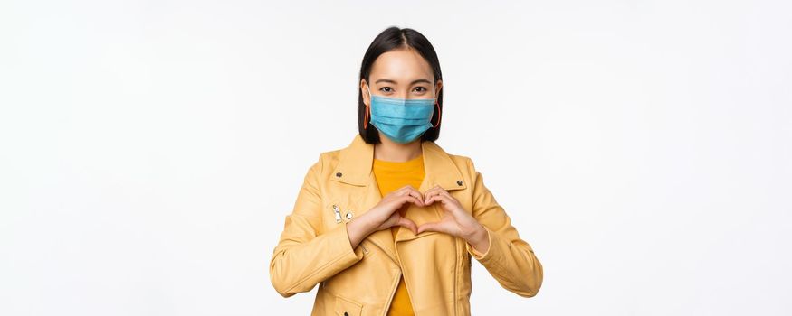 Covid-19 and people concept. Young asian woman in medical face mask, showing heart gesture and smiling with care, white background.