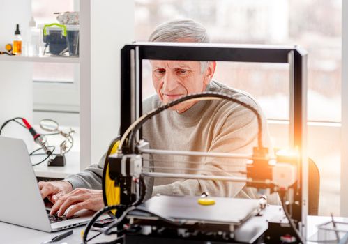 Adult wise man typing on laptop and looking at modern 3D printer in labaratory
