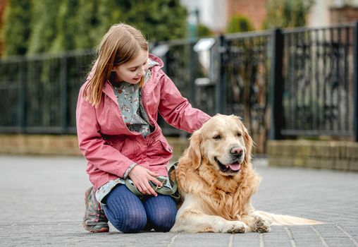 Preteen girl petting golden retriever dog at street. Female child kid with purebred dog labrador sits on ground.