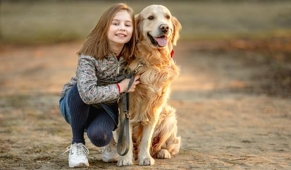 Preteen girl with golden retriever dog sitting in park in beautiful spring autumn day. Adorable female child kid hugging doggy pet outdoors and smiling portrait