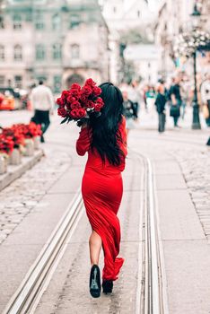 Elegant beautiful brunette woman in red fashion dress walking on the street , holding big bouquet of red roses. Birthday. March 8. Valentines day. obsession with red.