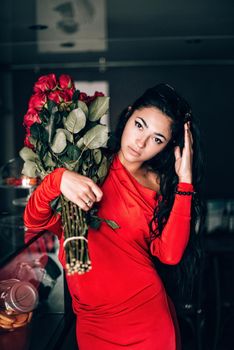Charming young woman in red sexy dress and sunglasses posing with a bouquet of red roses. photo of a seductive woman with black hair. Selective focus, filmgrain.