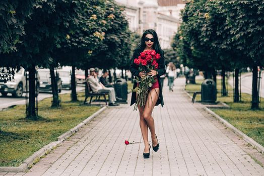 Elegant beautiful brunette woman in red shorts and black coat posing on a city streets with a big bouquet of red roses. Birthday. Valentines day.
