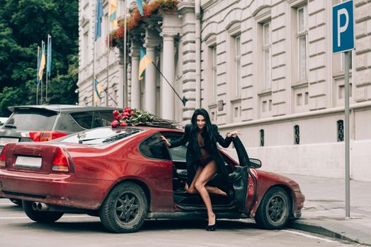 Elegant beautiful brunette woman in red shorts and black coat posing near red car. big bouquet of red roses on a car roof. Birthday. March 8.