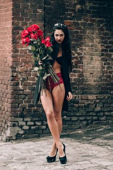Elegant beautiful brunette woman in red shorts and black coat posing near brick wall with a big bouquet of red roses. Birthday. March 8. Valentines day.