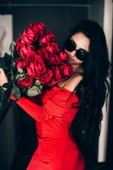 Charming young woman in red sexy dress and sunglasses posing with a bouquet of red roses. photo of a seductive woman with black hair. Selective focus, filmgrain.